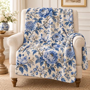 Puede incluir: Una manta de color crema con un patrón repetitivo de flores azul oscuro, ramas con hojas y pequeños pájaros. La manta está colocada sobre un sillón tapizado, mostrando el intrincado diseño botánico en tonos de azul.
