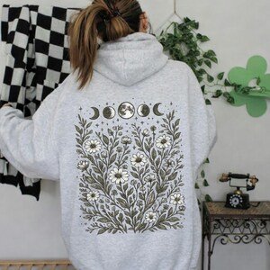 May include: Light gray hoodie with a floral and moon phase design on the back. The design features white flowers, green leaves, and the phases of the moon. The word "Ash" is printed in the bottom right corner.
