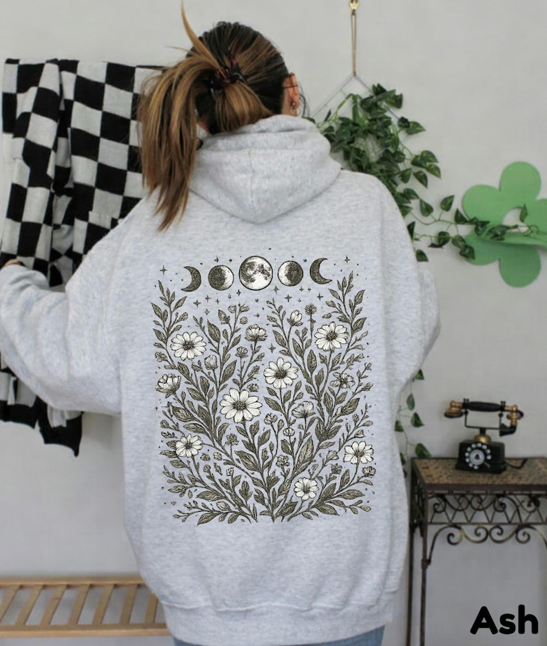 May include: Light gray hoodie with a floral and moon phase design on the back. The design features white flowers, green leaves, and the phases of the moon. The word "Ash" is printed in the bottom right corner.