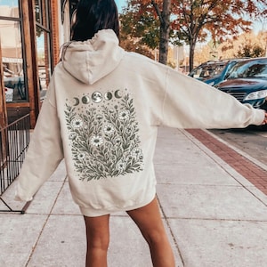 Puede incluir: Sudadera con capucha color arena con un diseño floral y de fases lunares en la espalda. El gráfico presenta flores blancas, hojas verdes y fases lunares. La palabra "Sand" está impresa en la parte inferior.