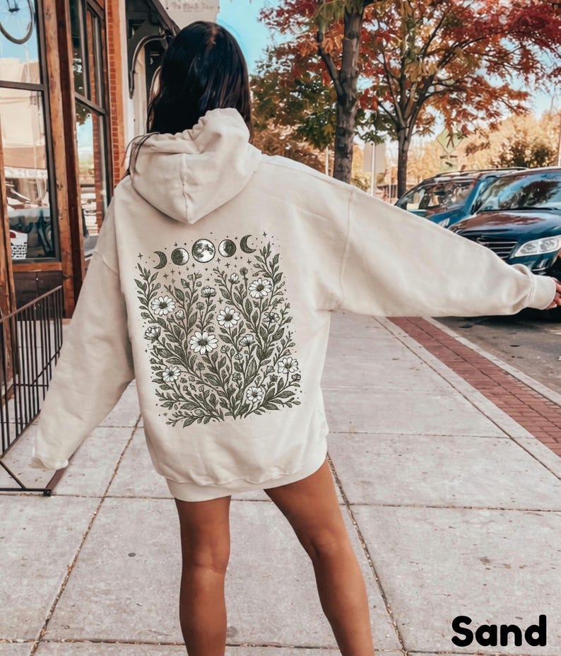 May include: Sand-colored hoodie with a floral and moon phase design on the back. The graphic features white flowers, green leaves, and moon phases. The word "Sand" is printed at the bottom.