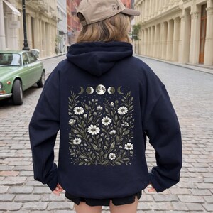 May include: Navy blue hoodie featuring a floral and moon phase design on the back. The design includes white daisies, green leaves, and a series of moon phases. The word "Navy" is printed at the bottom of the image.