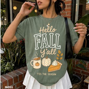 May include: A sage green t-shirt featuring the text Hello FALL Y'ALL TIS THE SEASON in white and orange lettering. The graphic includes a mug with a pumpkin design, a white pumpkin, and a slice of pumpkin pie with whipped cream on top.