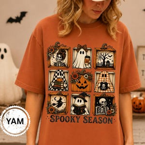 May include: An orange t-shirt featuring a grid of nine square illustrations including ghosts, skeletons, a jack-o'-lantern, and a witch. The graphic includes the text "SPOOKY SEASON" in a dark, distressed font at the bottom of the design.