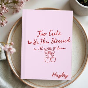Op de afbeelding: Een roze notitieboek met de tekst "Too Cute to Be This Stressed, so I'll write it down." in rode letters. Het notitieboek heeft een kersenillustratie en de naam "Hayley" onderaan. Het notitieboek ligt op een decoratieve schaal.