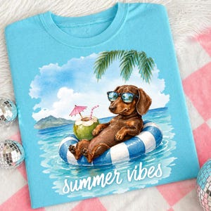May include: Pale blue t-shirt with a dachshund wearing sunglasses, floating in a blue and white striped inflatable ring in the sea. A coconut drink with a pink parasol is beside it. The words "summer vibes" are written in white.