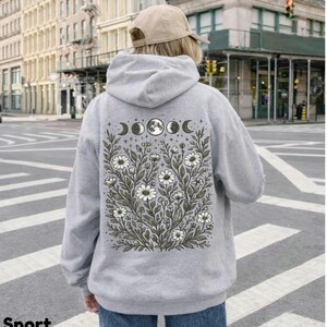 May include: Sport grey hoodie with a floral and moon phase design on the back. The design features white flowers, green leaves, and moon phases. The hoodie is a light grey color and has a hood and long sleeves. The text "Sport Grey" is at the bottom left.