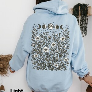 May include: Light blue hoodie with a floral and moon phase design on the back. The graphic features white daisies, leaves, and the phases of the moon. The text "Light Blue" is at the bottom left.