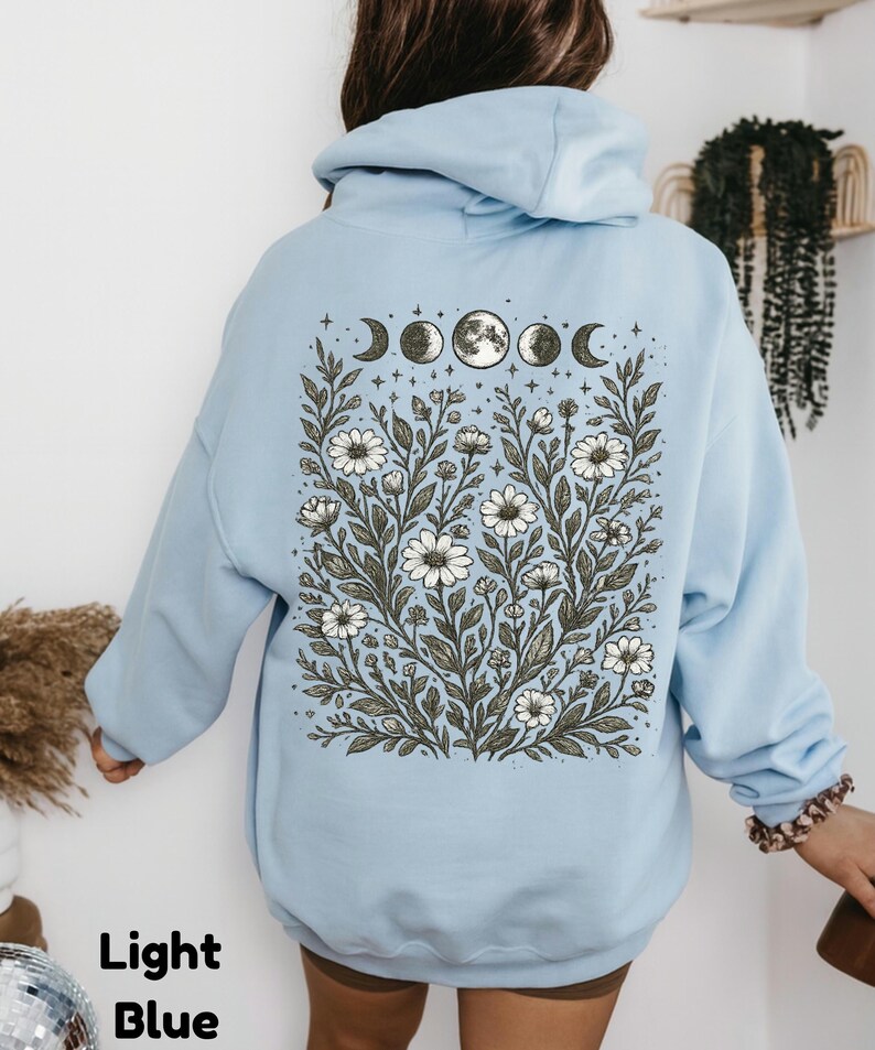 May include: Light blue hoodie with a floral and moon phase design on the back. The graphic features white daisies, leaves, and the phases of the moon. The text "Light Blue" is at the bottom left.
