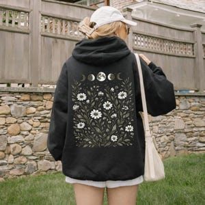 May include: Black hoodie with a floral and moon phase design on the back. The design features white daisies and moon phases. The word "Black" is printed at the bottom of the image. The hoodie is worn by a person wearing a white cap and sunglasses.