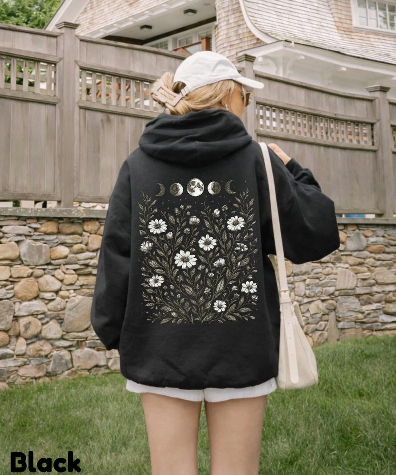 May include: Black hoodie with a floral and moon phase design on the back. The design features white daisies and moon phases. The word "Black" is printed at the bottom of the image. The hoodie is worn by a person wearing a white cap and sunglasses.