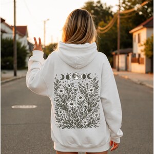 May include: White hoodie with a floral and moon phase design on the back. The graphic features daisies, stars, and crescent moons. The word "White" is printed at the bottom of the image.