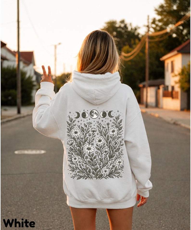 May include: White hoodie with a floral and moon phase design on the back. The graphic features daisies, stars, and crescent moons. The word "White" is printed at the bottom of the image.