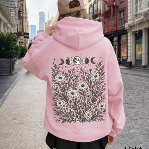 May include: Light pink hoodie with a floral and moon phase design on the back. The graphic features white daisies, leaves, and the phases of the moon. The text "Light Pink" is at the bottom right.