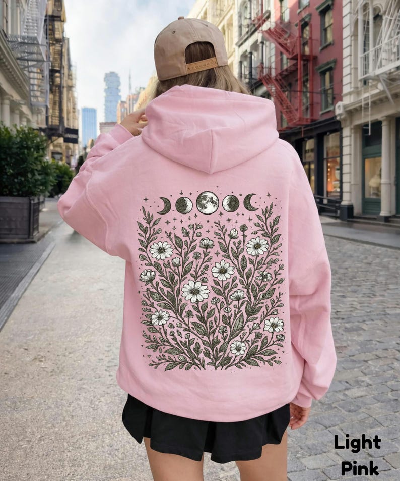 May include: Light pink hoodie with a floral and moon phase design on the back. The graphic features white daisies, leaves, and the phases of the moon. The text "Light Pink" is at the bottom right.