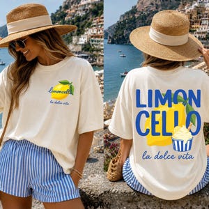 May include: Cream-colored t-shirt with a lemon graphic and the text "Limoncello la dolce vita." The shirt is paired with blue and white striped shorts and a straw hat. The back of the shirt has a larger lemon graphic and the text "LIMONCELLO la dolce vita."