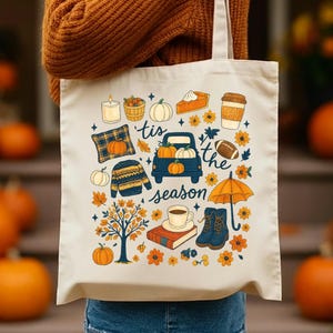 May include: Cream-colored tote bag featuring a fall-themed design. The design includes pumpkins, a blue truck carrying pumpkins, a coffee cup, a football, and the text "'tis the season".