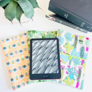 May include: A black Kindle e-reader with a screen displaying a pencil pattern, positioned over three fabric sleeves. The sleeves feature colorful succulent and cactus prints in shades of peach, green, blue, and pink, each with a small snap button.