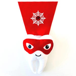 May include: A white plush tooth with a red superhero mask and cape. The red cape features a white spiderweb design with a black spider in the center. The tooth has a smiling face and is designed for children.