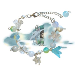 May include: A delicate bracelet featuring a mix of beads in white, blue, and green. The bracelet includes flower and star charms, with a silver chain and clasp. The background features an illustration of a dragon and a girl.