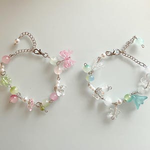 May include: Two delicate bracelets with silver chains, adorned with a variety of beads in pastel pink, green, and white. One bracelet features a pink butterfly charm, while the other has a blue flower charm. Both have a pearl at the end of the chain.