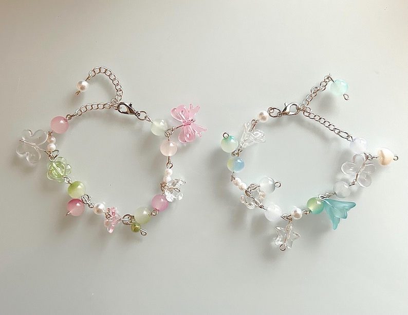 May include: Two delicate bracelets with silver chains, adorned with a variety of beads in pastel pink, green, and white. One bracelet features a pink butterfly charm, while the other has a blue flower charm. Both have a pearl at the end of the chain.