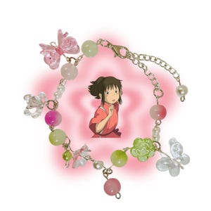 May include: A delicate bracelet featuring a variety of beads in pink, green, and white, along with clear butterfly and flower charms. The bracelet has a gold-colored chain and clasp. A cartoon image of a girl is in the background.