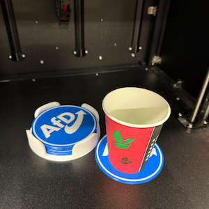 Może przedstawiać: Dwa okrągłe podkładki pod kubki, jeden niebieski z tekstem "AFD" i białym znacznikiem wyboru, a drugi niebieski z czerwono-zielonym wzorem. Biały papierowy kubek z czerwono-zielonym wzorem stoi na drugim podkładce.