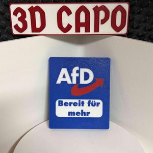 Könnte beinhalten: Ein rechteckiges Schild mit dem Text "3D CAPO" in roten Buchstaben auf weißem Hintergrund und ein blaues Quadrat mit den Buchstaben "AfD" und den Worten "Bereit für mehr" in Weiß.