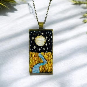 Puede incluir: Un collar con cadena dorada y un colgante rectangular con una escena bordada de una luna llena, estrellas y un río que fluye a través de un campo de trigo amarillo.
