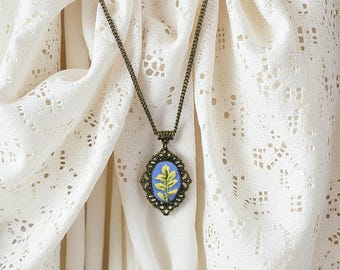 May include: A vintage necklace with a bronze chain and pendant. The pendant has an oval design with a blue background and a yellow and green embroidered leaf. The necklace is set against a white lace background, creating a delicate aesthetic.
