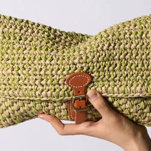 Könnte beinhalten: Eine gewebte Clutch in Grün- und Beigetönen, gehalten von einer Hand. Die Tasche hat ein strukturiertes Muster und einen braunen Lederriemen mit Metallschnalle. Die Clutch ist etwa 30 cm breit.