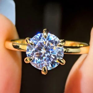May include: A gold ring featuring a large, clear, round-cut diamond. The diamond is set in a six-prong setting.