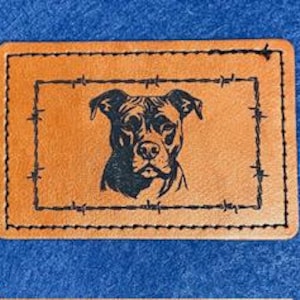 May include: A brown leather patch with a black stitched border and a black silhouette of a pit bull dog inside a barbed wire frame.