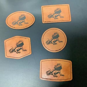 Peut inclure: Cinq patchs en cuir marron de différentes formes : ovale, rectangle, cercle et bouclier. Chaque patch présente une illustration noire d'un dragon barbu. Les patchs sont cousus sur les bords.