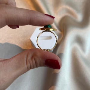 Puede incluir: Un anillo dorado con una banda retorcida y una piedra verde circular. El anillo se sostiene entre dos dedos sobre un fondo blanco y una tela sedosa de color blanco roto.
