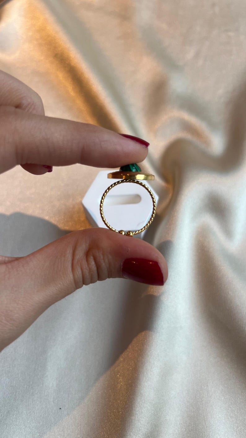 Puede incluir: Un anillo dorado con una banda retorcida y una piedra verde circular. El anillo se sostiene entre dos dedos sobre un fondo blanco y una tela sedosa de color blanco roto.