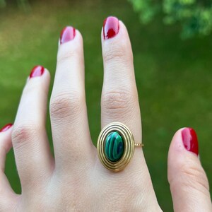 Puede incluir: Anillo de oro con una piedra ovalada de malaquita con dise&ntilde;o de c&iacute;rculos conc&eacute;ntricos. El anillo est&aacute; engastado en una fina banda de oro. El fondo es verde borroso.