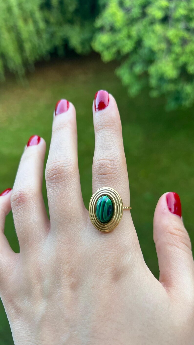 Puede incluir: Anillo de oro con una piedra ovalada de malaquita con dise&ntilde;o de c&iacute;rculos conc&eacute;ntricos. El anillo est&aacute; engastado en una fina banda de oro. El fondo es verde borroso.