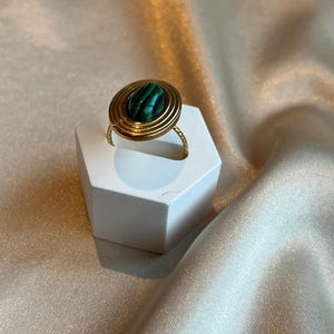 Puede incluir: Un anillo dorado con una piedra de malaquita. El anillo presenta un dise&ntilde;o en espiral alrededor de la piedra y una banda retorcida. La piedra de malaquita es de forma ovalada con rayas verdes y negras. El anillo se exhibe sobre un soporte hexagonal blanco.