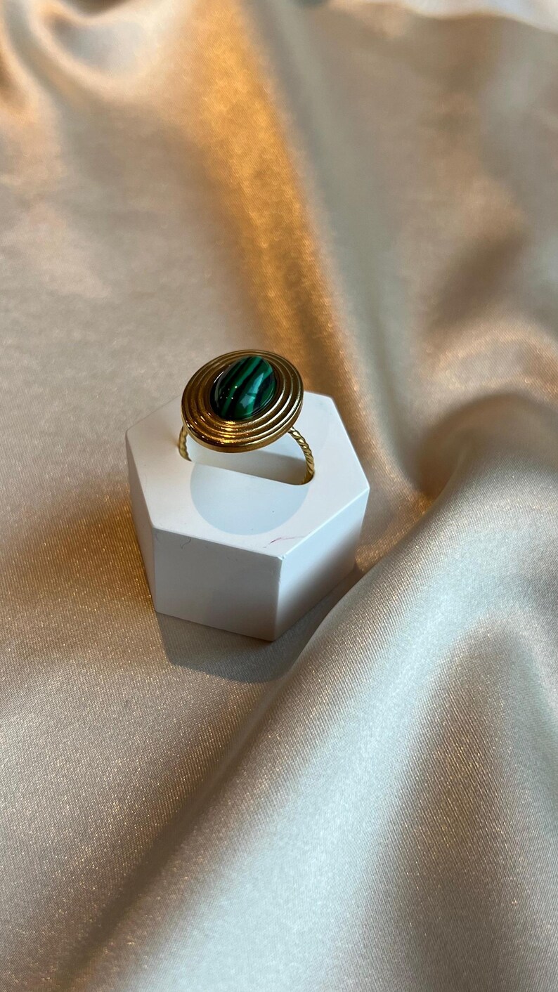 Puede incluir: Un anillo dorado con una piedra de malaquita. El anillo presenta un dise&ntilde;o en espiral alrededor de la piedra y una banda retorcida. La piedra de malaquita es de forma ovalada con rayas verdes y negras. El anillo se exhibe sobre un soporte hexagonal blanco.