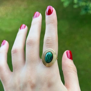 Puede incluir: Anillo dorado con una piedra ovalada de malaquita con patrones verdes y negros. El anillo tiene un dise&ntilde;o en espiral alrededor de la piedra. El fondo es verde borroso, lo que sugiere un entorno al aire libre. Las u&ntilde;as est&aacute;n pintadas de rojo.