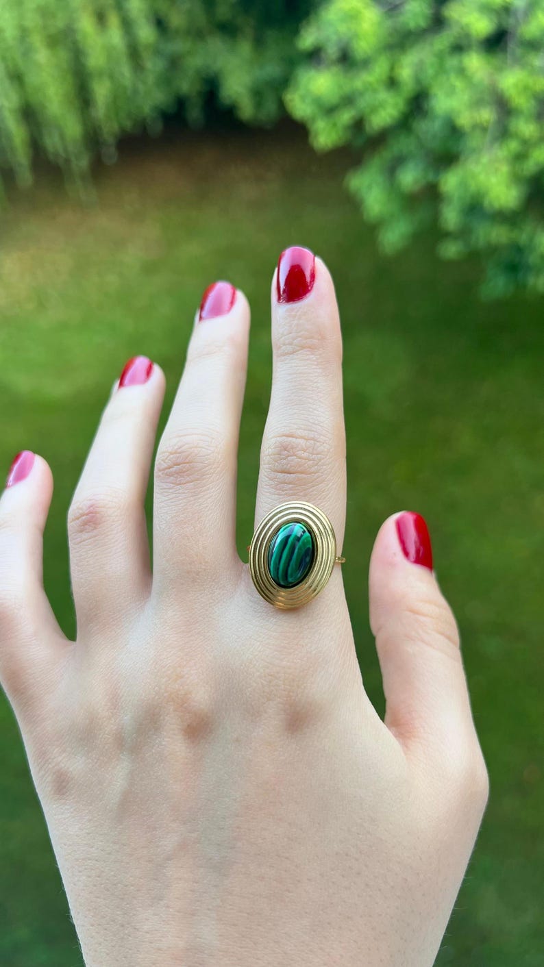 Puede incluir: Anillo dorado con una piedra ovalada de malaquita con patrones verdes y negros. El anillo tiene un dise&ntilde;o en espiral alrededor de la piedra. El fondo es verde borroso, lo que sugiere un entorno al aire libre. Las u&ntilde;as est&aacute;n pintadas de rojo.