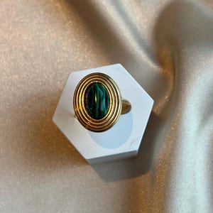 Puede incluir: Un anillo dorado con una piedra de malaquita. El anillo presenta una banda dorada y un engaste circular con crestas conc&eacute;ntricas alrededor de la piedra ovalada de color verde oscuro. El anillo se exhibe sobre un soporte hexagonal blanco sobre un fondo satinado.