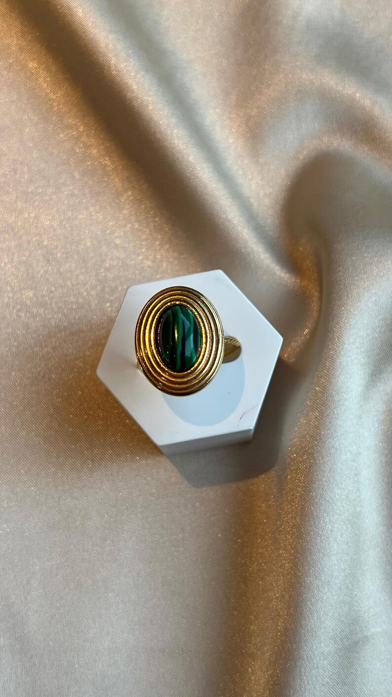 Puede incluir: Un anillo dorado con una piedra de malaquita. El anillo presenta una banda dorada y un engaste circular con crestas conc&eacute;ntricas alrededor de la piedra ovalada de color verde oscuro. El anillo se exhibe sobre un soporte hexagonal blanco sobre un fondo satinado.