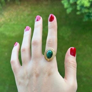 Puede incluir: Un anillo dorado con una piedra verde ovalada. El anillo tiene un dise&ntilde;o en capas alrededor de la piedra. Las u&ntilde;as est&aacute;n pintadas de rojo. El fondo es verde borroso.