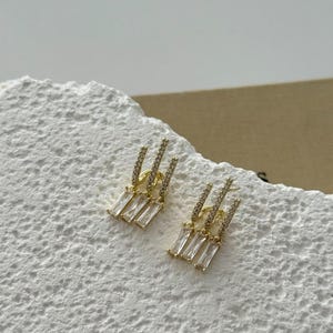 May include: A pair of gold-coloured earrings with a dangling design. Each earring features three rectangular, clear crystals suspended below a vertical row of smaller, sparkling crystals. The earrings are displayed on a textured, white surface.