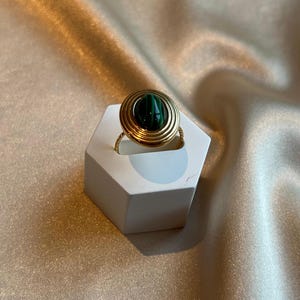 Puede incluir: Un anillo dorado con una piedra de malaquita engastada en un dise&ntilde;o en espiral. El anillo se exhibe sobre un soporte hexagonal blanco, sobre un fondo de tela satinada.