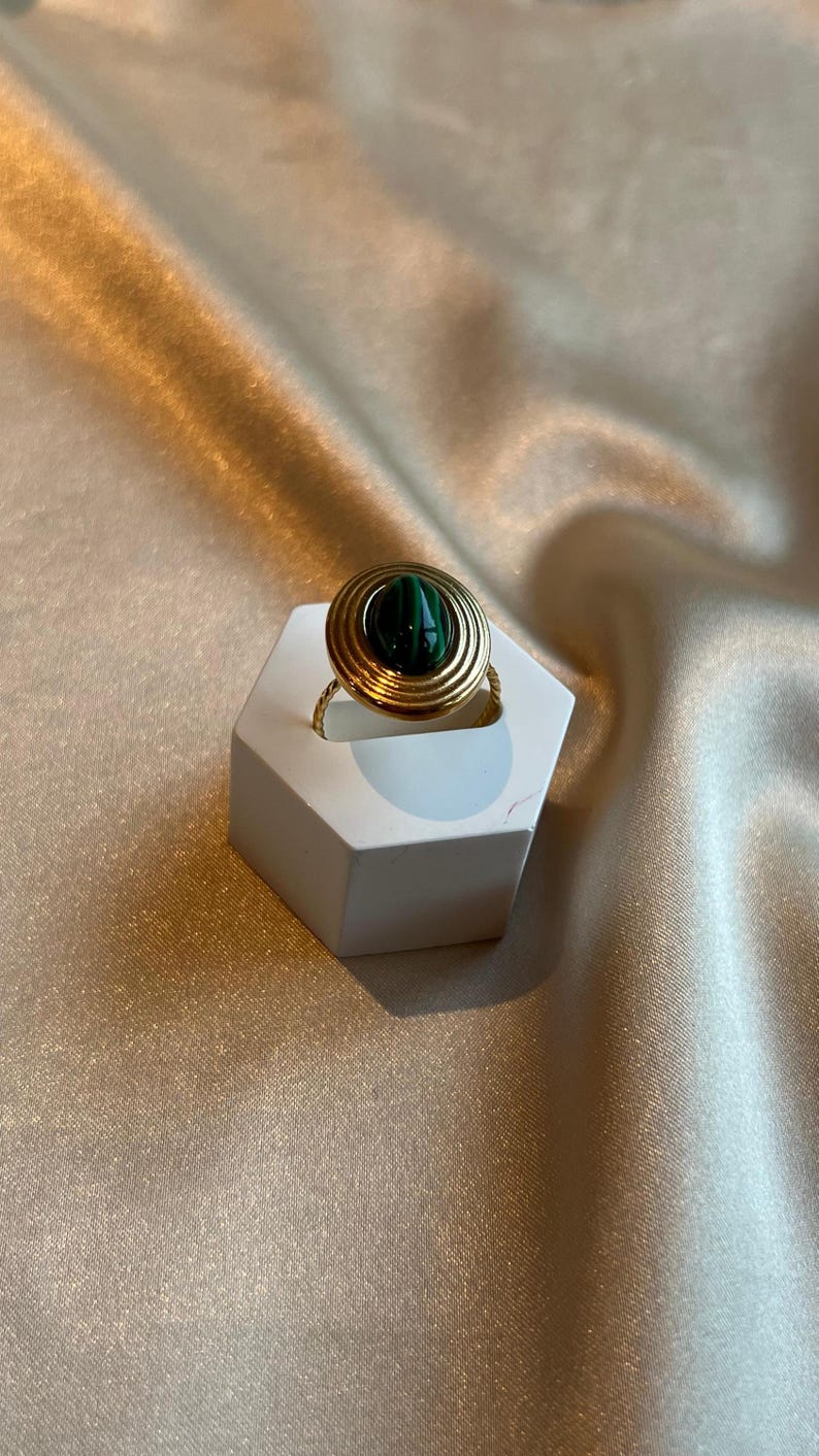 Puede incluir: Un anillo dorado con una piedra de malaquita engastada en un dise&ntilde;o en espiral. El anillo se exhibe sobre un soporte hexagonal blanco, sobre un fondo de tela satinada.