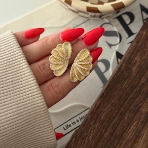 May include: A pair of gold-colored, fan-shaped earrings with intricate detailing and small, clear stones. The earrings are held in a hand with red nail polish. The phrase "Life is a journey" is visible in the background.